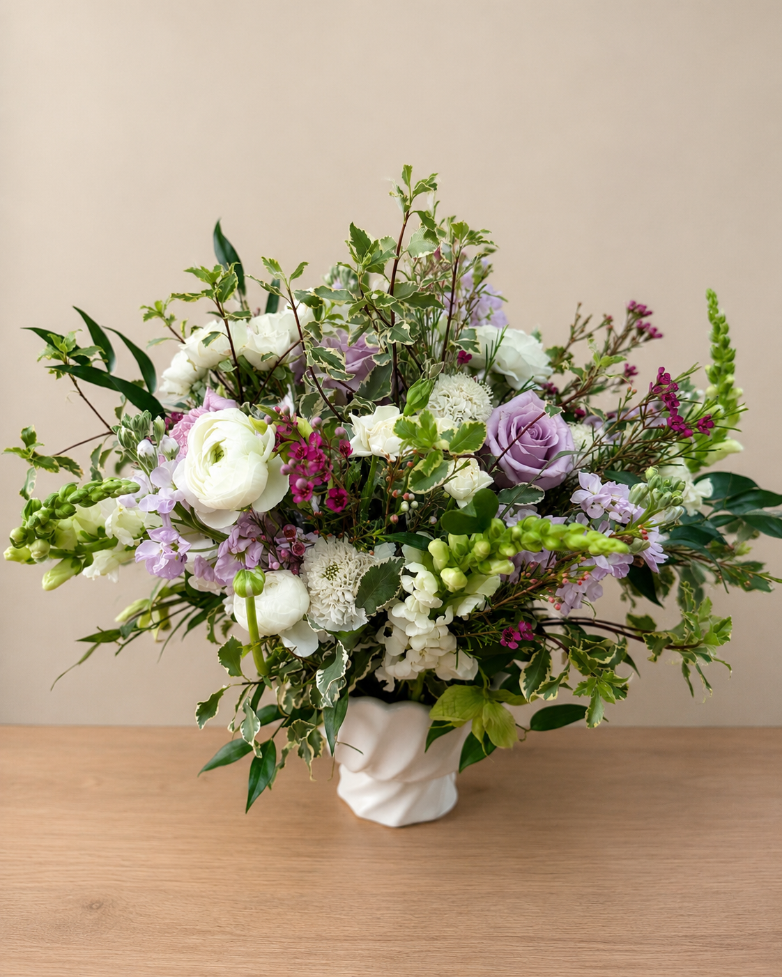 Garden style arrangement in purple and white with ranunculus, rose, snap dragon, stock, scabiosa, spray rose in a white compote vase.